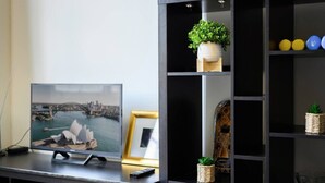 Una habitación con un televisor sobre un mueble bajo, un cuadro de un puente y un estante negro con objetos decorativos.