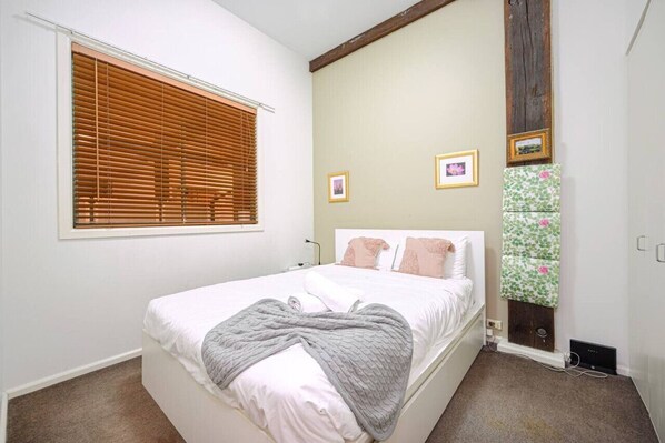 A bedroom with a bed, a window with blinds, a wall-mounted heater, and framed pictures. - Sydney CBD 1Br with Spa/Pool/Sauna/Gym by ICC (Pyrmont)