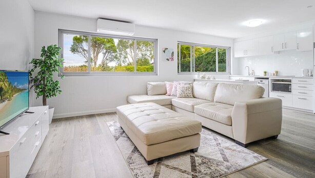 A modern living room with a sectional sofa, a flat-screen TV, and a large window with a view of trees. - Collaroy 3Br with pool / Breath and Relax (Collaroy)