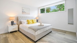 A bedroom with a large bed, white walls, a window with a view of greenery, and a bedside table with a lamp. - Collaroy 3Br with pool / Breath and Relax (Collaroy)