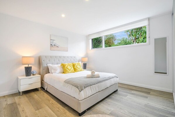 A bedroom with a large bed, white walls, a window with a view of greenery, and a bedside table with a lamp. - Collaroy 3Br with pool / Breath and Relax (Collaroy)
