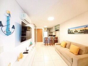 A living room with a sofa, a TV, a kitchen area with bar stools, and a painting on the wall. - Carneiros Beach Resort by CTA (Tamandaré)