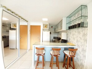 A kitchen with a central island, three wooden bar stools, a refrigerator, a microwave, and a glass cabinet with black cups. - Carneiros Beach Resort by CTA (Tamandaré)