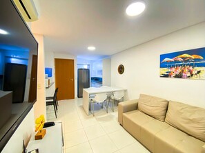 Una sala de estar con un sofá color crema, una mesa de comedor con sillas, una cocina al fondo y un cuadro en la pared.