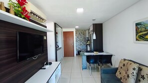 A modern living room with a flat-screen TV, a sofa, a dining area with a table and chairs, and a kitchen in the background. - Carneiros Beach Resort by CTA (Tamandaré)