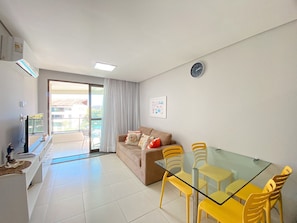 A modern living room with a glass dining table, yellow chairs, a sofa with cushions, a TV, and a sliding glass door leading to a balcony. - Carneiros Beach Resort by CTA (Tamandaré)