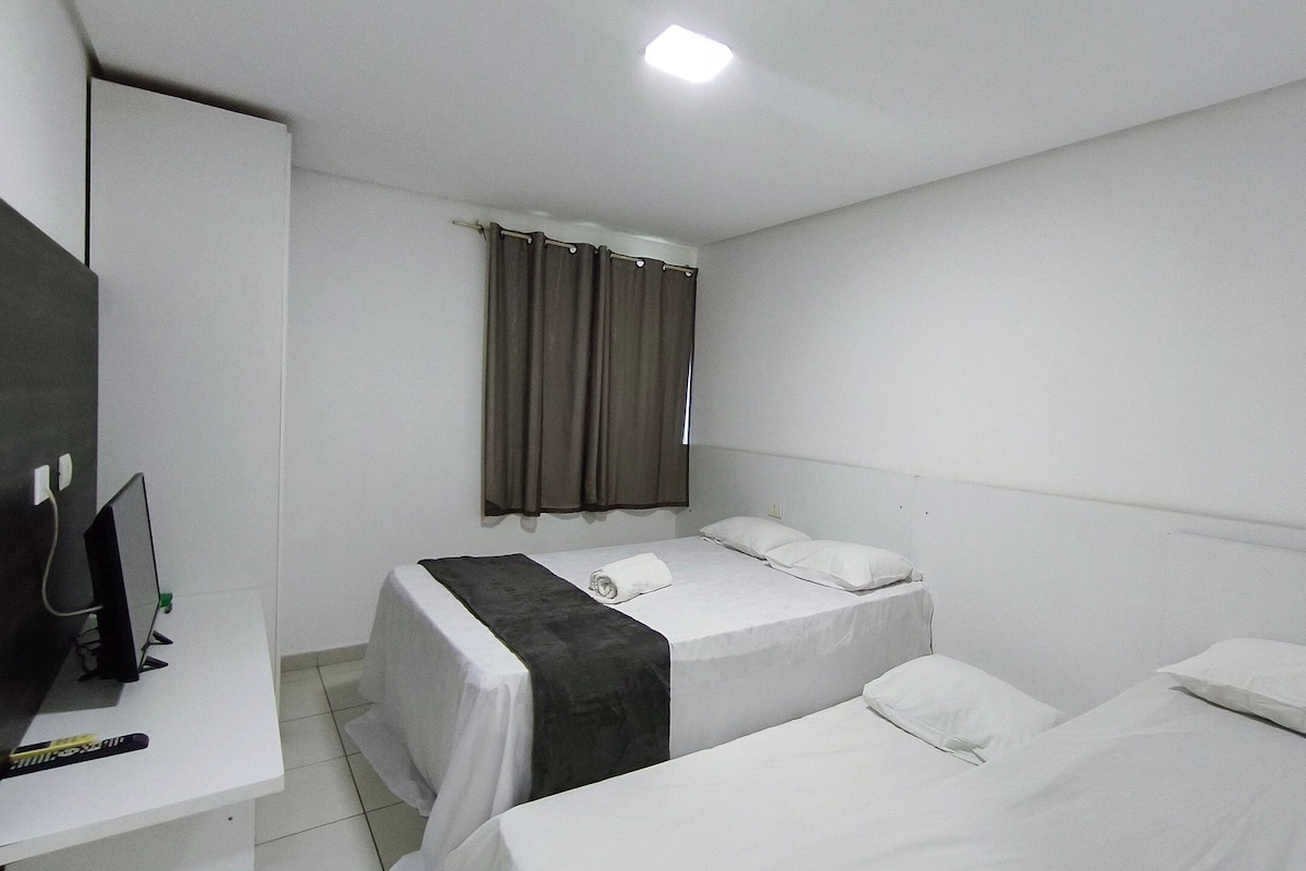 Habitación de hotel con dos camas individuales, un escritorio con silla, televisión y ventana con cortinas.