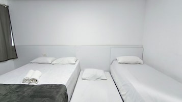 Una habitación con dos camas individuales, cada una con ropa de cama y almohadas blancas, una manta gris en la cama de la izquierda, y una ventana con cortina al lado izquierdo.