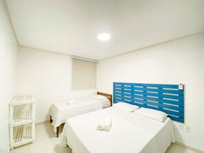 Habitación de hotel con dos camas, una ventana con persianas, un estante blanco y un cabecero azul.