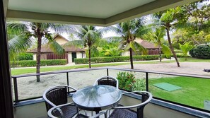 A balcony with a glass table and chairs, overlooking a well-maintained garden with palm trees and a building in the background. - Carneiros Beach Resort by CTA (Tamandaré)