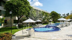 A pool area with lounge chairs and umbrellas, surrounded by greenery and a building. - Carneiros Beach Resort by CTA (Tamandaré)