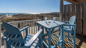 A wooden deck with blue Adirondack chairs and a table, overlooking a beach and dunes. - Oceanfront Condo, Beach Access, Shared Pool, & Hot Tub (Hatteras)
