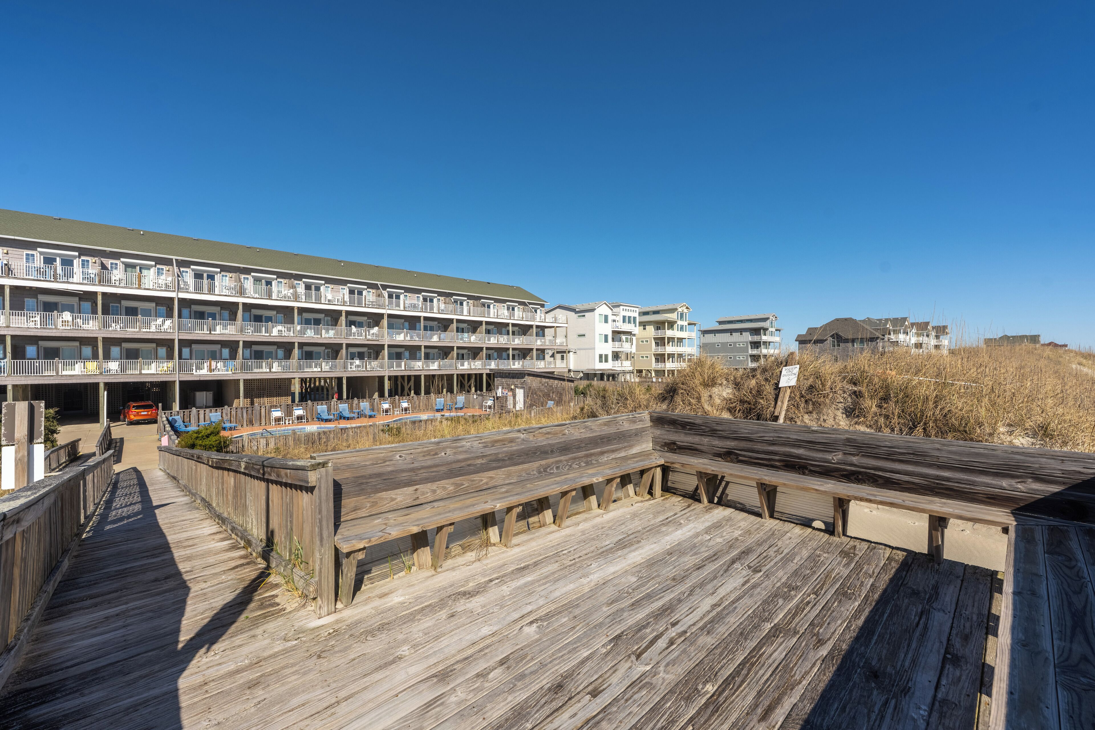 Un muelle de madera que conduce a un hotel frente al mar, con varios balcones y una zona de piscina.