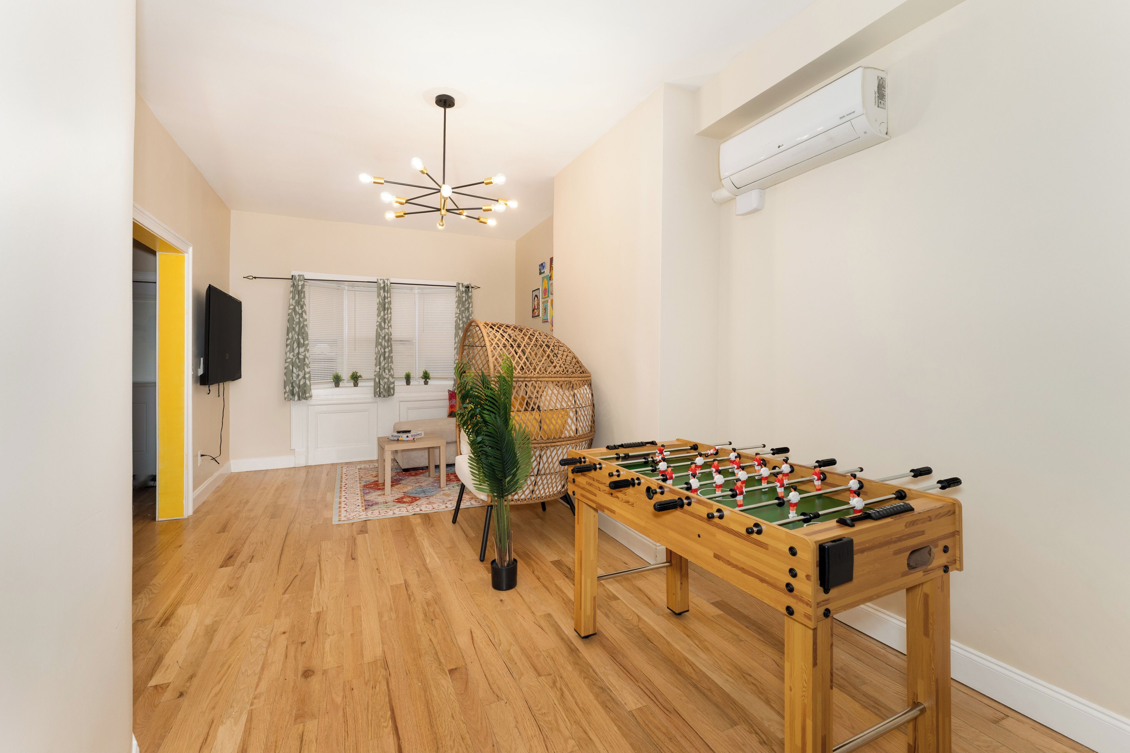 Una habitación con una mesa de fútbol de madera, una silla de mimbre, una pequeña mesa con una planta y una lámpara en el techo.
