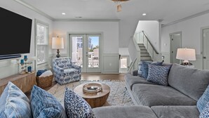 Un salon spacieux avec un grand canapé gris, un fauteuil à motifs et une table basse en bois. Un ventilateur de plafond et un téléviseur sont fixés au mur.