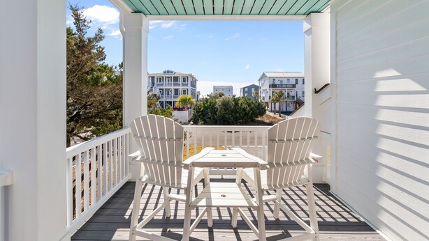 Un balcon abrité, doté de chaises blanches et d’une table, qui offre une vue sur un quartier résidentiel avec ses maisons et ses arbres.