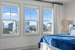 A bedroom with a large bed, three windows, and a view of houses.
