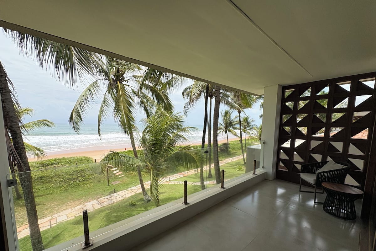 A balcony with a view of the beach, palm trees, and a modern interior with a geometric wall design and seating.