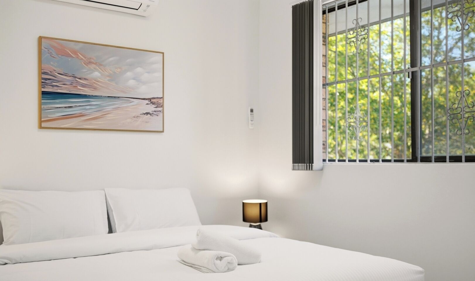 A hotel room with a bed, a painting, a window with bars, and a lamp.