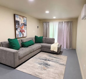 Una sala de estar con un sofá seccional gris, cojines verdes, un cuadro abstracto grande, una manta color beige y una alfombra gris.