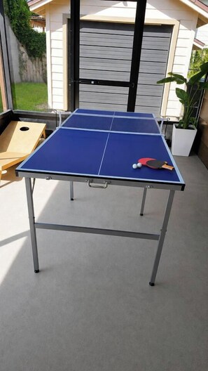 Una mesa de ping pong con una paleta y una pelota, montada al aire libre con una casa al fondo.