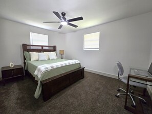 A bedroom with a bed, a desk with a chair, a ceiling fan, and two windows with blinds. - Historic Avondale 5BR | Porch, Grill & Parking (Jacksonville)