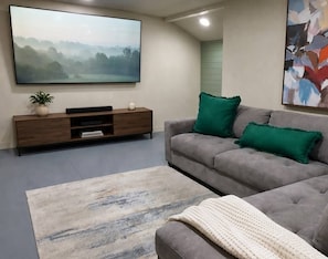 Un salon avec un canapé gris, un téléviseur à écran plat, un meuble de rangement en bois et un grand tableau abstrait.