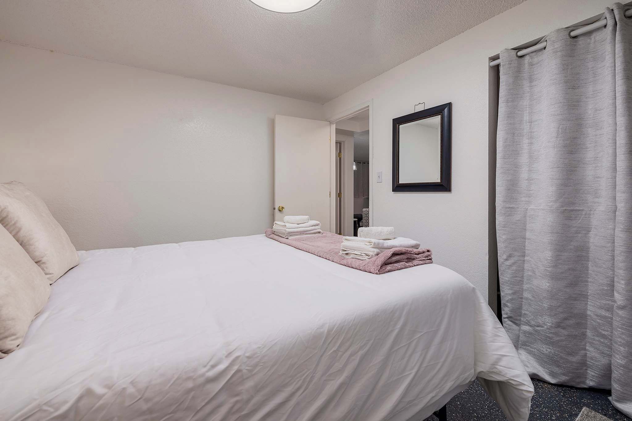 Ein ordentlich bezogenes Bett mit weißen Laken und einer rosa Decke, ein Spiegel an der Wand und ein grauer Vorhang.