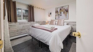 Un dormitorio con cama, ventana con cortinas, mesita de noche con lámpara y un cuadro en la pared.