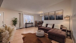 A modern living room with a brown sofa, a round coffee table, and a cowhide rug. There is a dining area with a table and chairs, and a large window with curtains. - Cozy South Addition Downtown Getaway - King Beds (Anchorage)