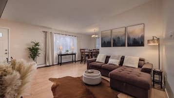 Un salon moderne avec un canapé marron, une table basse ronde et un tapis en peau de vache. On y trouve également un coin repas avec une table et des chaises, ainsi qu’une grande fenêtre avec des rideaux.