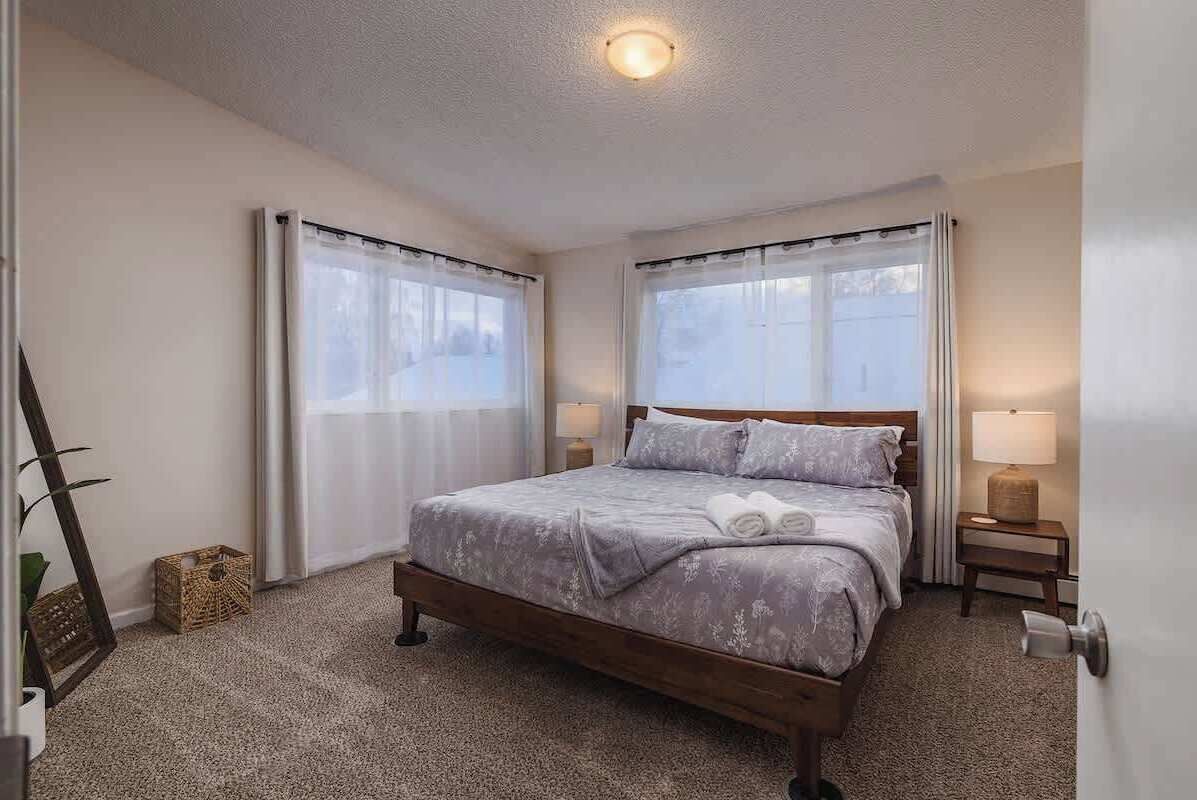 Une chambre à coucher bien rangée, avec un grand lit, deux tables de chevet avec des lampes et une fenêtre avec des rideaux.
