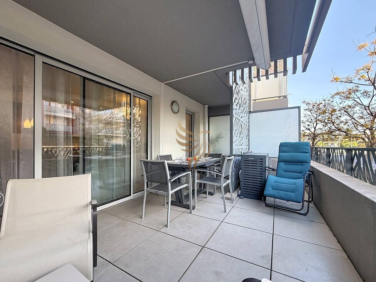 Un balcon avec une table et des chaises pour prendre les repas, un fauteuil bleu et une porte vitrée donnant sur une pièce.