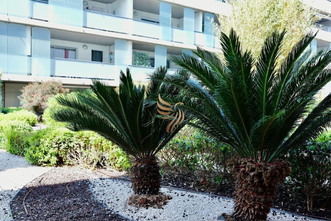 A modern building with multiple balconies and a well-maintained garden with palm trees.