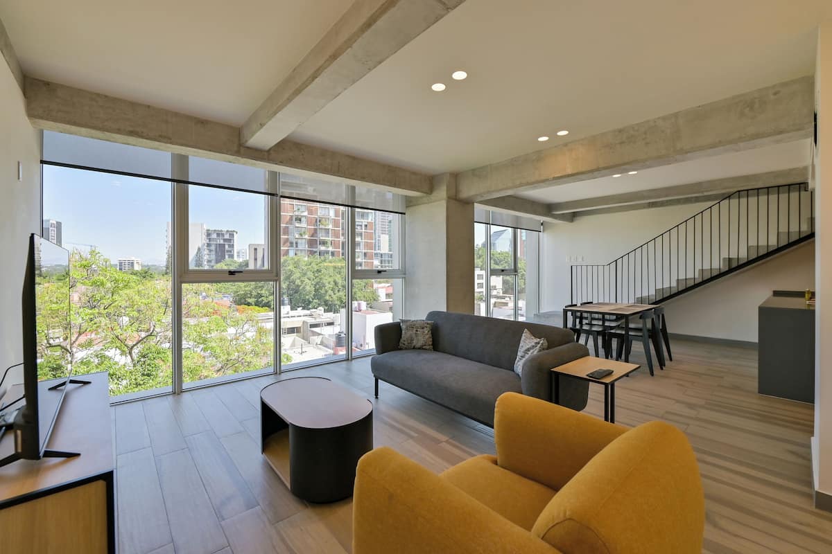 Un salon moderne avec un canapé, un fauteuil jaune, une table basse et un téléviseur. Un escalier mène à un niveau supérieur.