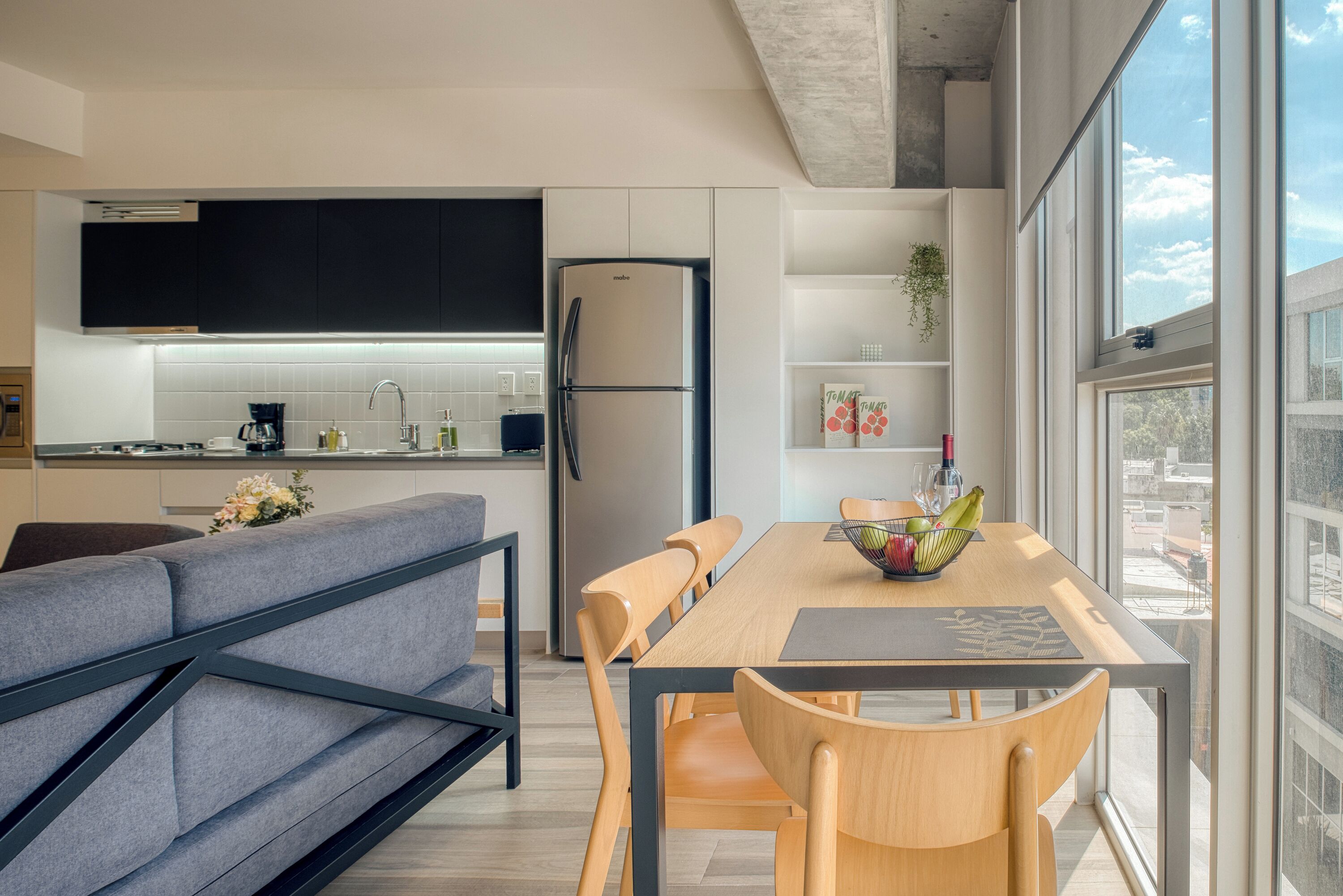 A modern kitchen with a built-in refrigerator, a dining area with a wooden table and chairs, and a large window offering a city view.