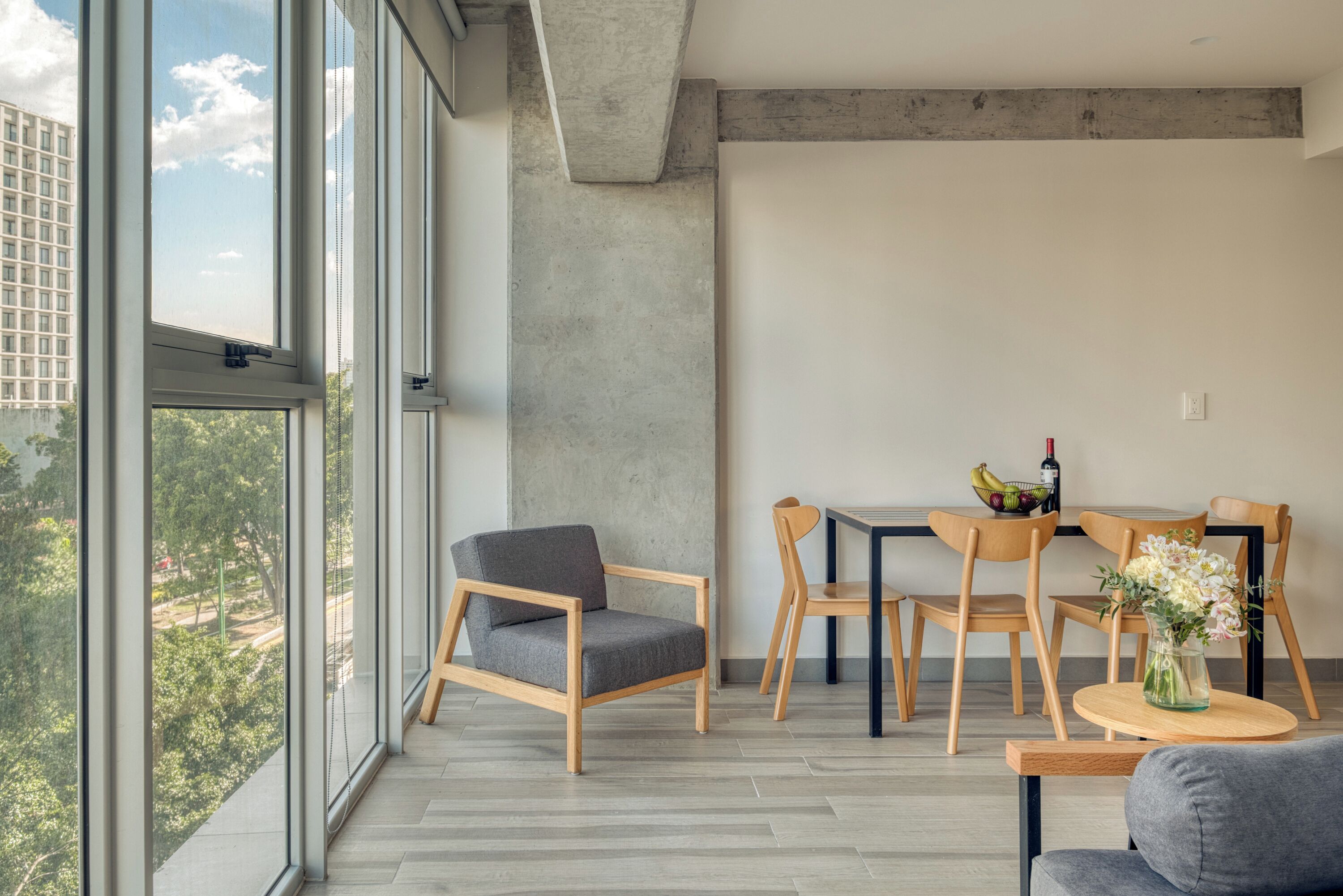 A modern living room with large windows, a dining area with wooden chairs and a table, and a grey sofa.