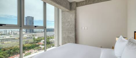 Una habitación de hotel moderna con una cama grande, vista de la ciudad a través de ventanas de piso a techo y una pared de acento de concreto.