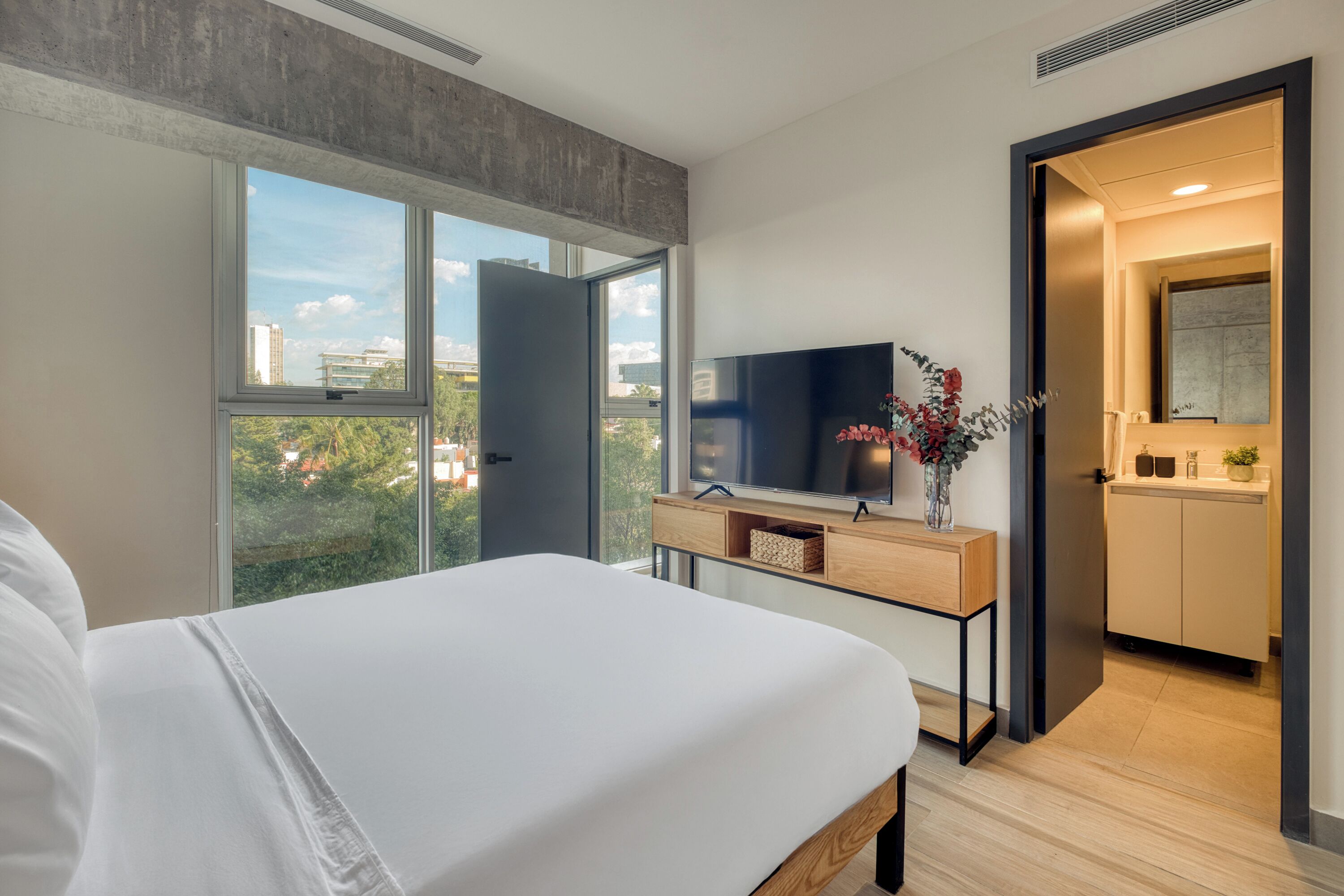 Habitación de hotel moderna con una cama grande, televisor, una mesa consola de madera y vistas de la ciudad a través de amplios ventanales.