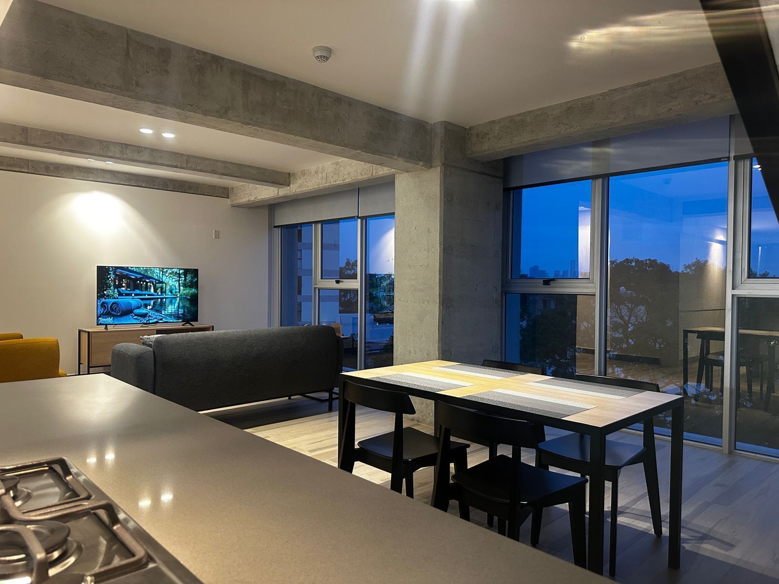 A modern kitchen with a dining area, large windows, and a flat-screen TV.