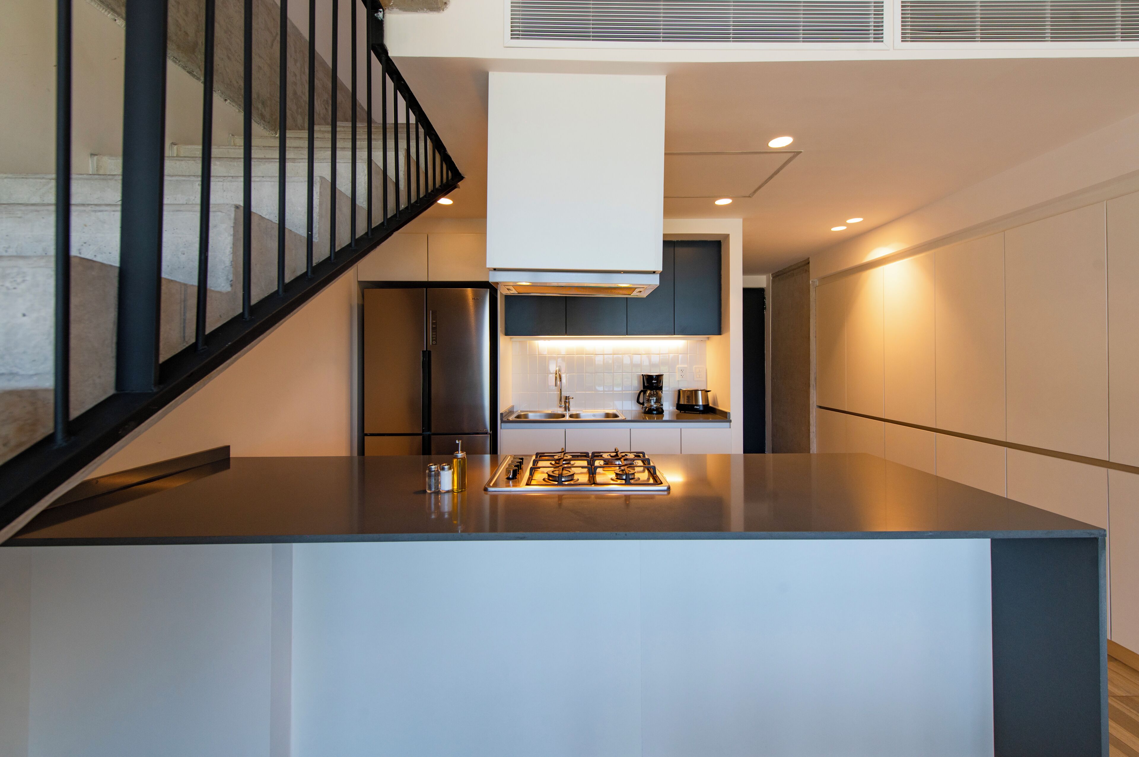 Une cuisine moderne dotée d’un îlot central, d’appareils électroménagers en acier inoxydable et d’un escalier avec une rampe noire.