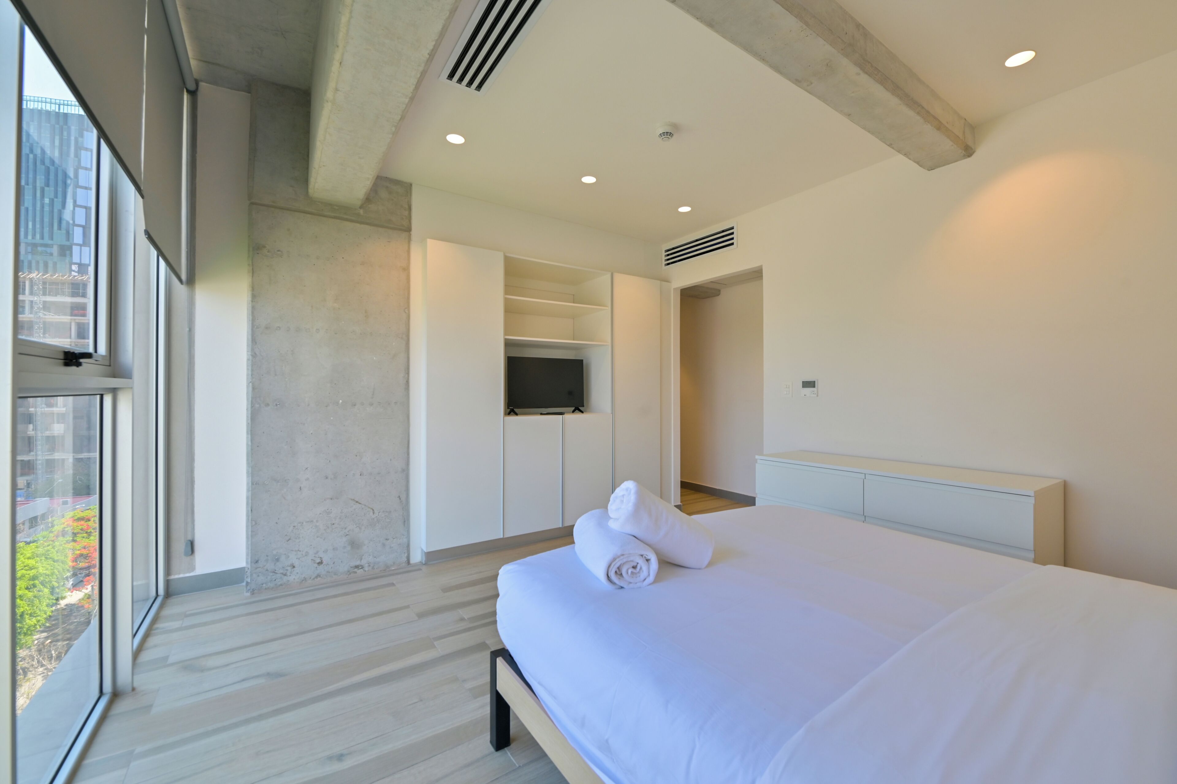 A modern hotel room with a large bed, a walk-in closet, a television, and a view of greenery outside.