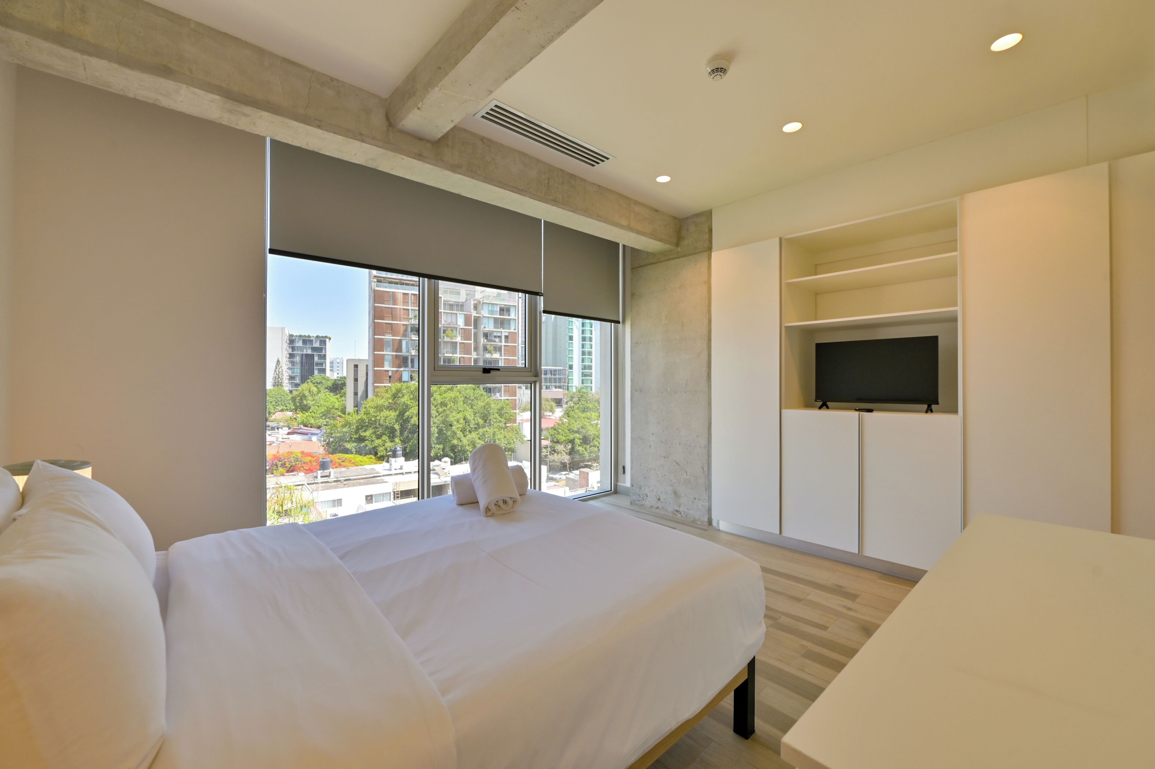 A modern hotel room with a large bed, a TV mounted on the wall, and a view of the cityscape through large windows.