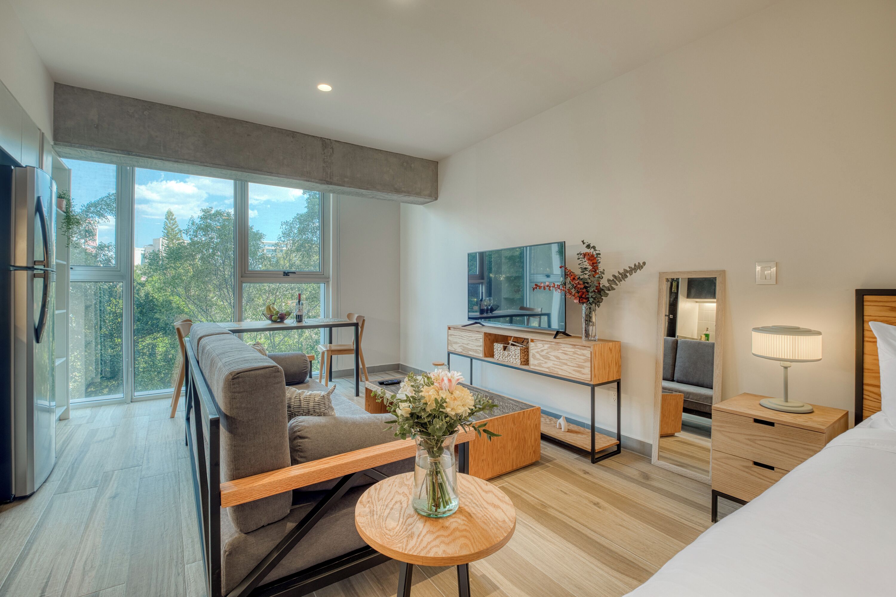 A modern hotel room with a sofa, a small dining table, a TV, a bedside table with a lamp, and a large window offering a view of trees.