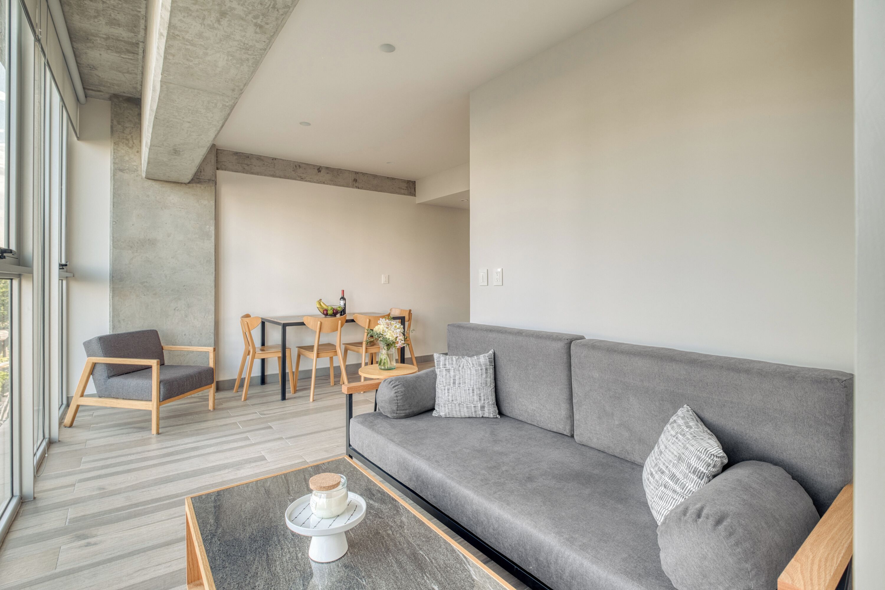 A modern living room with a grey sofa, wooden chairs, a coffee table, and a dining area with a table and chairs.