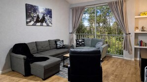 Ein modernes Wohnzimmer mit einem grauen Sofa, einem schwarzen Sessel und einem Gemälde einer Straßenszene. Ein großes Fenster mit Vorhängen bietet Blick auf Bäume.
