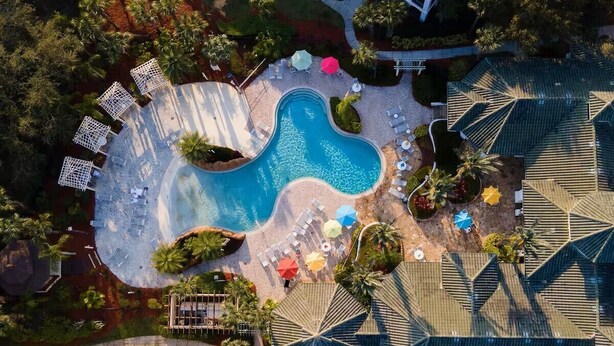 Eine Vogelperspektive eines Resorts mit einem zentralen Schwimmbecken, Liegestühlen, Sonnenschirmen und Palmen.