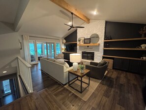 A modern living room with a fireplace, a ceiling fan, and a large window overlooking a deck.