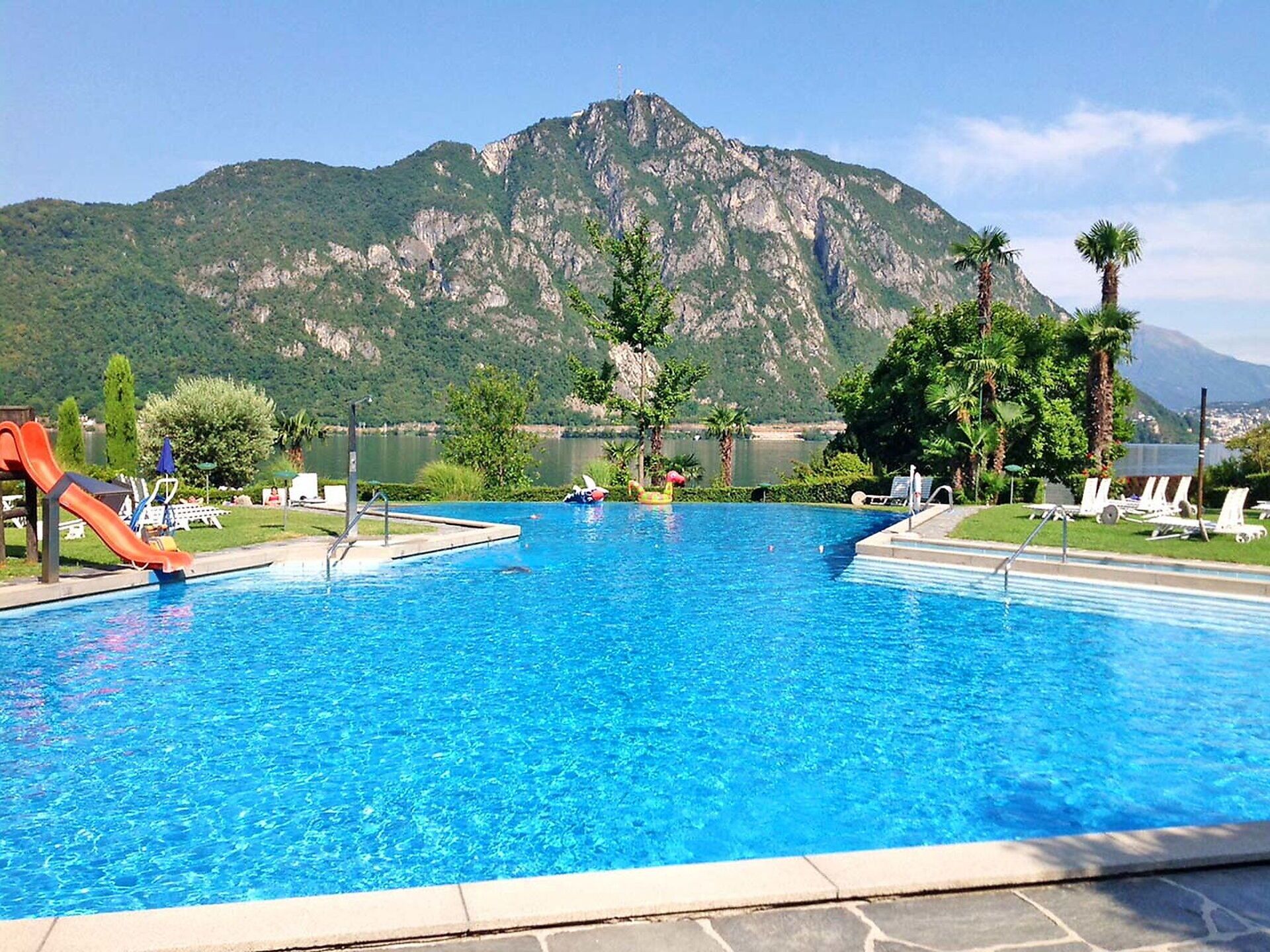 A swimming pool with a mountain backdrop, surrounded by palm trees and lounge chairs.