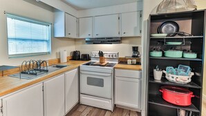 Une cuisine compacte avec des meubles blancs, un plan de travail en bois, une cuisinière, un évier et une étagère contenant des ustensiles de cuisine.